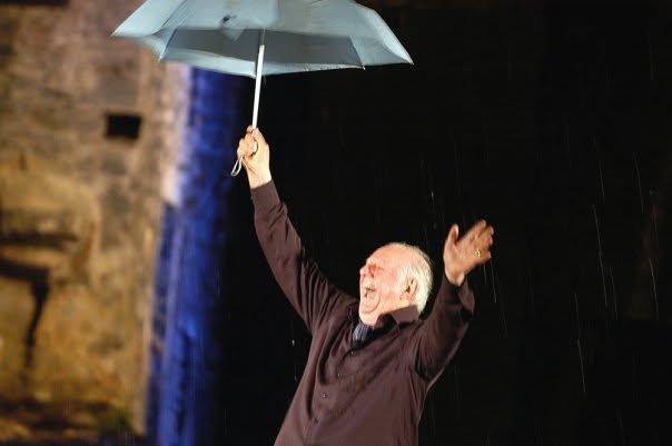 Mistero Buffo, a performer with an umbrella in the rain, monastery in the background