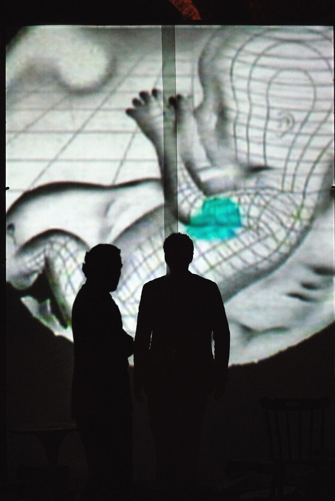A Number, two silhouetted figures before a projected anatomical backdrop